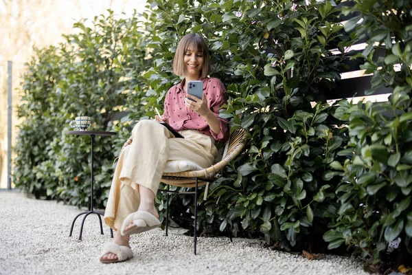 Young cheerful woman works on smart phone and tablet while sitting on background of green bushes outdoors. Concept of remote work at cozy garden