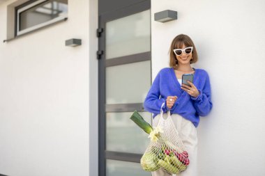 Şık bir kadın evinin verandasında elinde bir çanta dolusu taze sebzeyle dikilirken telefonu kullanıyor. Sürdürülebilirlik, sağlıklı gıda ve modern çevre dostu yaşam tarzı kavramı