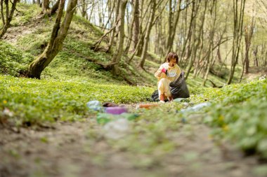 Koruyucu elbiseli genç bir kadın ormanda dağınık plastik çöpleri topluyor. Yatak ekolojisi ve çevre kirliliği sorunu uzun zamandır çürümekte olan atıkla