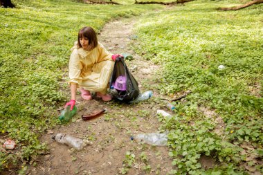 Koruyucu elbiseli genç bir kadın ormanda dağınık plastik çöpleri topluyor. Yatak ekolojisi ve çevre kirliliği sorunu uzun zamandır çürümekte olan atıkla