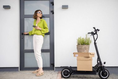 Evinin verandasında elektrikli scooter ve paketlerle duran bir kadın. Sürdürülebilirlik ve modern çevre dostu yaşam tarzı kavramı