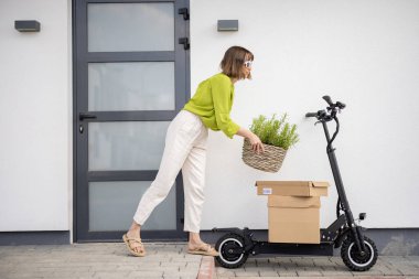 Evinin verandasında yanında elektrikli scooter ve paketlerle ot taşıyan genç bir kadın. Sürdürülebilirlik kavramı ve modern çevre dostu yaşam tarzı