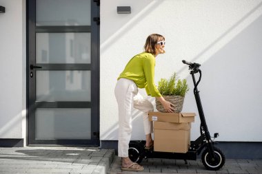Evinin verandasında yanında elektrikli scooter ve paketlerle ot taşıyan genç bir kadın. Sürdürülebilirlik kavramı ve modern çevre dostu yaşam tarzı