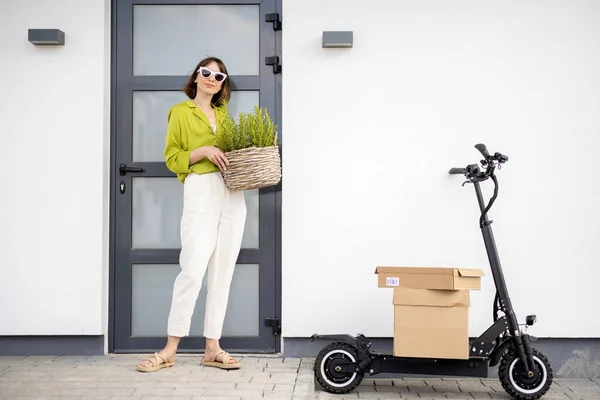 Young woman carrying basket with herbs on a porch of her house with electric scooter and parcels nearby. Concept of sustainability and modern eco-friendly lifestyle
