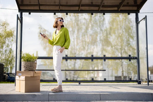 Young Woman Stands Parcels Flowerpot Bus Stop Using Phone Concept — Stockfoto