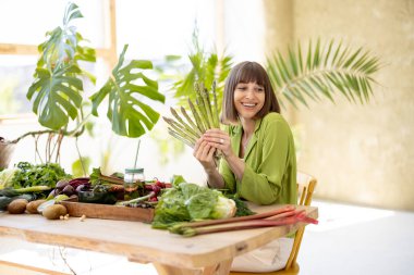 Genç ve neşeli bir kadının portresi taze sebze, meyve ve sebze dolu masada otururken bir sürü kuşkonmaz tutuyor. Sağlıklı beslenme ve yaşam tarzı kavramı