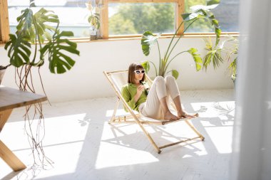 Kadın, güneşli oturma odasında arka planda yeşil bitkilerle güneşlenirken sağlıklı kase yiyor. Rahatlama ve sağlıklı yeme konsepti