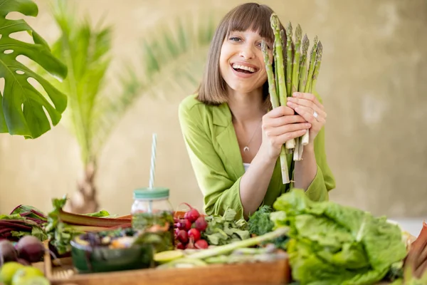 一个年轻而快乐的女人坐在餐桌旁 手里拿着一束芦笋 桌上有新鲜的蔬菜 水果和蔬菜 健康饮食和生活方式概念 — 图库照片
