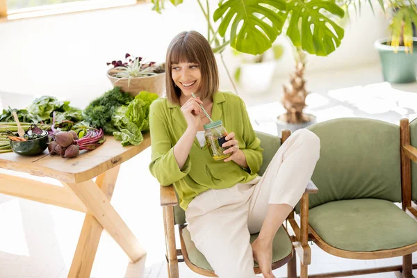 年轻的女人坐在靠近桌子的椅子上喝柠檬水 在房间里放着大量的新鲜食物配料和绿色植物 健康生活方式概念 — 图库照片