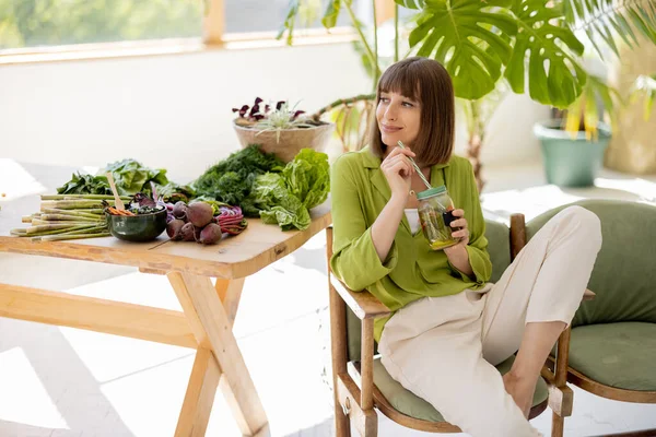 年轻的女人坐在靠近桌子的椅子上喝柠檬水 在房间里放着大量的新鲜食物配料和绿色植物 健康生活方式概念 — 图库照片