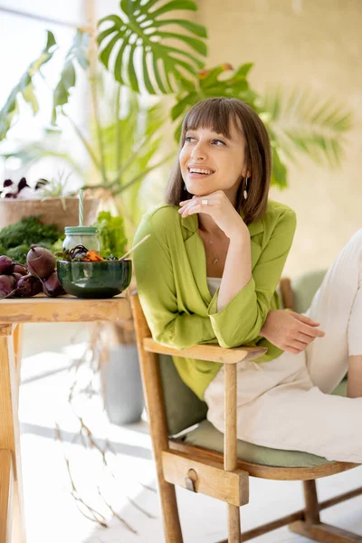 若い陽気な女性の肖像画は 背景に緑の植物と部屋の新鮮なビーガン料理でいっぱいのテーブルの近くの椅子にリラックス座っています 健康的な食事と健康の概念 — ストック写真