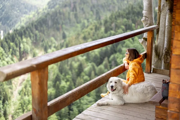 オレンジ色のスポーツスーツを着た若い人は 木製のテラスで彼女の犬と座っている間 素晴らしい山の風景を楽しんでいます 自然の中での脱出と孤独の概念とペットと一緒に旅行 — ストック写真