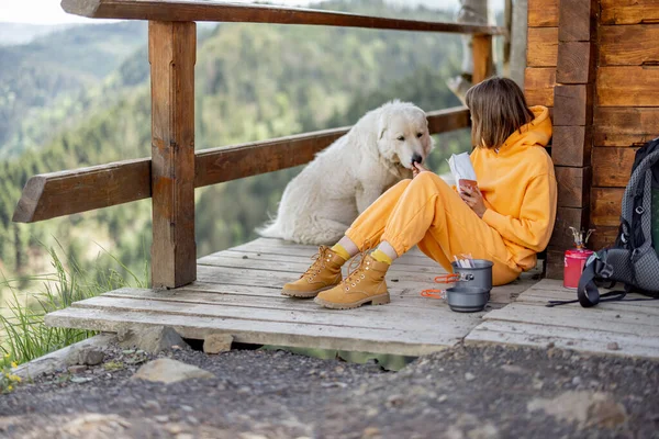 年轻女子与她的狗一起坐在一座木制的自然屋阳台上 一边欣赏高山美景 一边吃升华的食物 一边徒步旅行 大自然中的孤独和与宠物同行的概念 — 图库照片