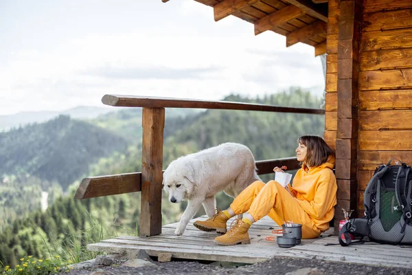 年轻女子与她的狗一起坐在一座木制的自然屋阳台上 一边欣赏高山美景 一边吃升华的食物 一边徒步旅行 大自然中的孤独和与宠物同行的概念 — 图库照片