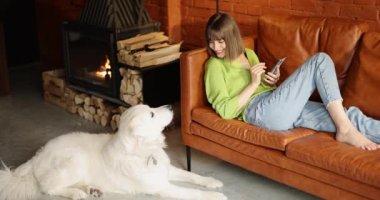 Kanepede tatlı beyaz köpeğiyle rahatça otururken telefonu kullanan genç bir kadın. Evcil hayvanlarla arkadaşlık ve evde boş zaman.