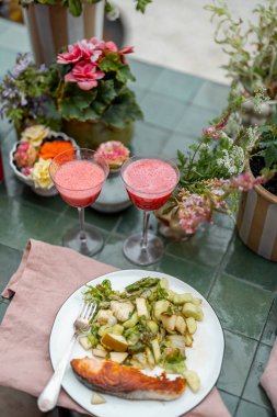 Açık havada çiçeklerle dolu bir masada öğle yemeği. Bahçedeki masada et ve kokteyl bulunan sebzeler.