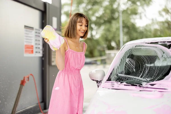 Young woman in pink dress wiping her tiny car covered in nano foam with a sponge at car wash. Concept of easy and beautiful self-service at car wash