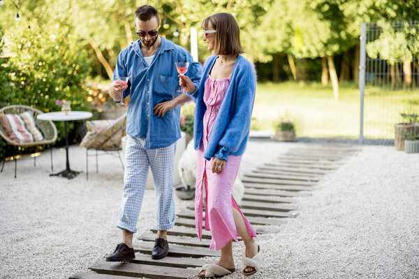 Young stylish couple have romantic conversation while walking with a drink at their beautiful backyard of country house. Young man and woman spend summer time happily