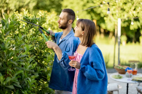 素敵なカップルは 自然の中で自分の国の家の裏庭で夏の時間を過ごします 緑の茂みを剪定する男 女性は飲み物を持っていて 夫と話す — ストック写真