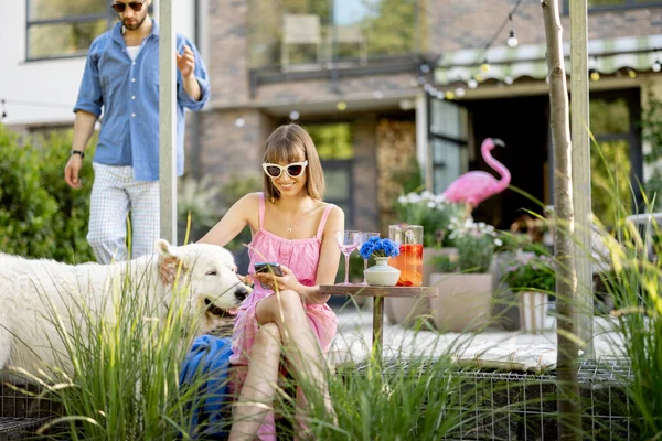 Stylish couple spend leisure time with a dog at beautiful backyard of their country house during summer time. Young happy family relax at their garden