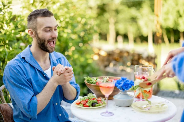 興奮した男が昼食を待っている 彼の妻は健康的なサラダを提供していますように熱狂的に見ている 若い家族は夏の間裏庭で昼食を食べます — ストック写真