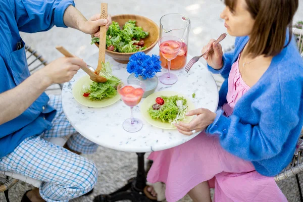 若く明るいカップルは 屋外の丸いテーブルに座っている間に健康的なサラダを食べ 夏の飲み物を飲みます 上からの眺め — ストック写真