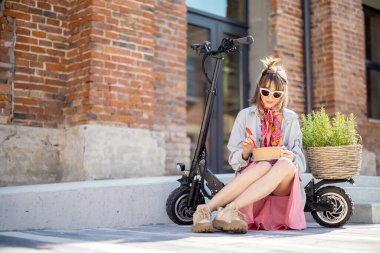 Genç ve şık bir kadın kahve molası veriyor, dışarıda yeşil bitkilerle elektrikli scooterın üzerinde otururken biraz yemek yiyor. Modern ve sürdürülebilir yaşam tarzı kavramı