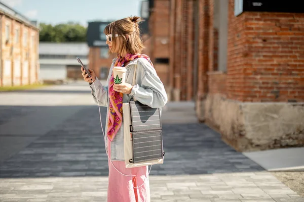 Young stylish woman walks with coffee and smart phone charging it from portable solar panel hanging on bag. Concept of modern and sustainable lifestyle