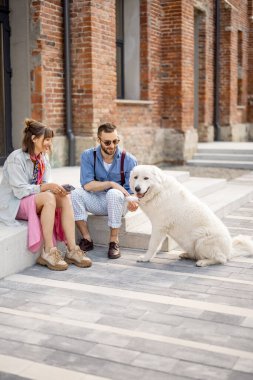 Genç ve şık bir çift sokakta beyaz köpekleriyle birlikte oturup eğleniyor. Genç hippiler ofisin yakınında birlikte takılıyor.