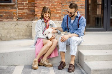 Genç ve şık bir çift sokakta beyaz köpekleriyle birlikte oturup eğleniyor. Genç hippiler ofisin yakınında birlikte takılıyor.