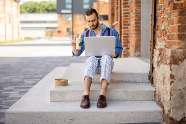一个时髦的家伙坐在笔记本电脑上 一边喝咖啡 一边在外面的街上吃东西 一边干活 现代商业生活方式和自由职业的概念 — 图库照片