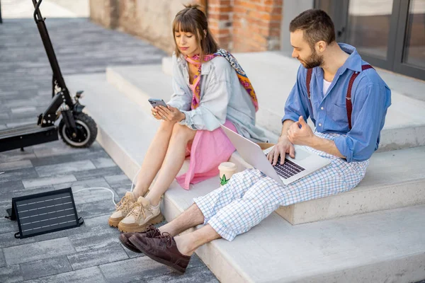 Stylish colleagues work on phone and laptop computer, charging them from portable solar panel, while sitting with electric scooter on a street at office district. Concept of sustainable lifestyle