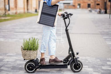 Adam elektrikli scooter kullanıyor ve hareket halindeyken telefonu güneş paneliyle şarj ediyor. Kesik manzara. Sürdürülebilir iş hayatı ve yeşil enerji kavramı