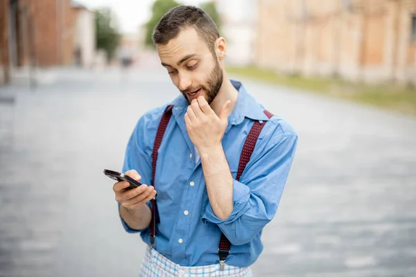 Portrait of weird businessman uses phone outdoors. Cool guy wearing blue shirt with suspenders