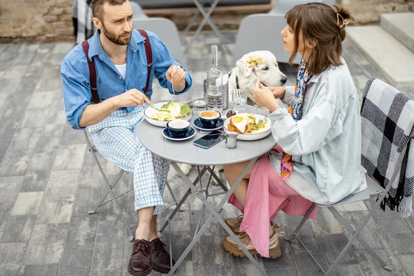 Stylish man and woman have delicious breakfast, sitting with cute dog at cafe terrace. Friends spending time together during a breakfast