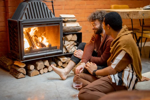 Two men talking while sitting together by the burning fireplace at cozy home. Concept of homosexual relations and coziness on winter time. Idea of multinational gay families