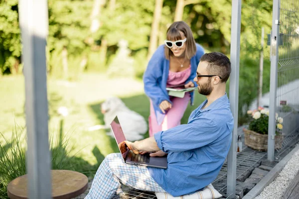 Young stylish couple work or spend leisure time with laptop computer on porch of country house on nature. Concept of remote work on nature