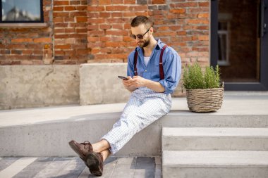 Stylish man uses smartphone while sitting on street with green plant near office building. Portrait of modern hipster with phone outdoors
