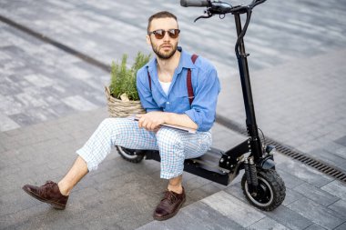 Portrait of stylish weird man sit with laptop on electric scooter with green plant on a street. Concept of sustainable and eco friendly lifestyle