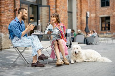 Modern ofis bölgesindeki kafe terasında köpekle otururken şık iki meslektaş sohbet ediyor.