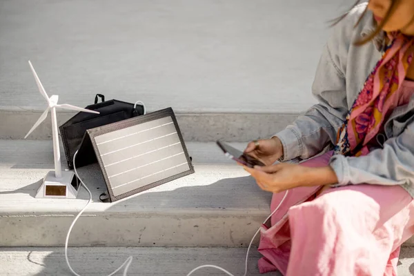 Person use smart phone and charge it from portable solar panel while sitting on street outdoors. Modern sustainable lifestyle and renewable energy concept