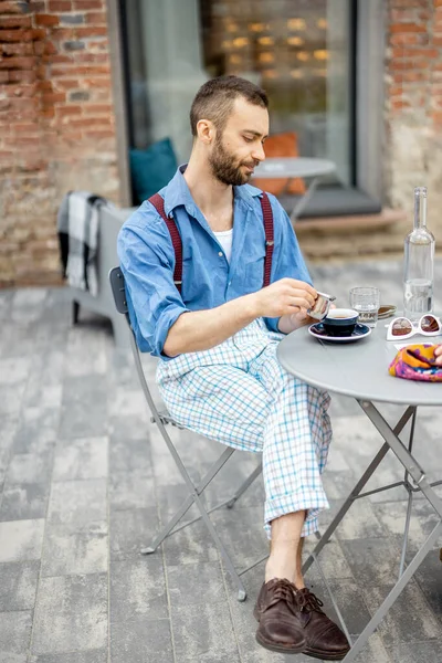 Stylish man drinks coffee while sitting at cafe outdoors. Leisure time. Guy wearing blue shirt with suspenders