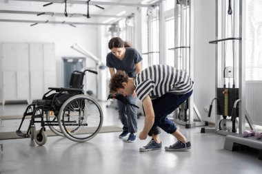 Guy wears cuffs on his legs in preparation for exercise, nurse with wheelchair on the background. Concept of physical therapy for people with disabilities