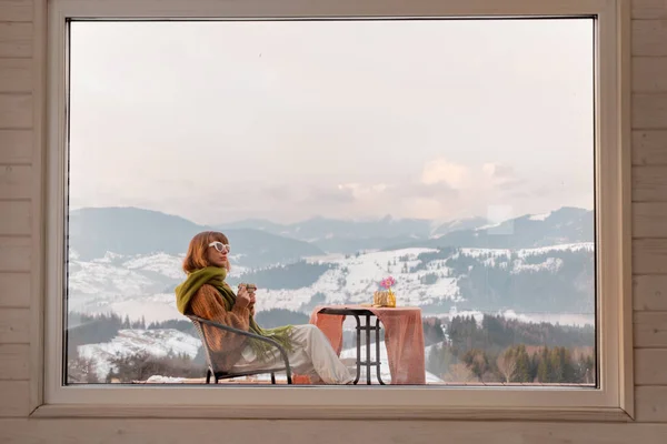 Woman sitting with a drink on terrace of tiny house in the mountains, view through the window from the inside. Concept of small modern cabins for rest and escape to nature