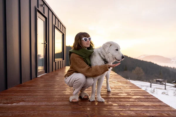 Woman hugs her dog while resting on terrace of tiny house in the mountains on winter time. Concept of small modern cabins for rest and escape to nature. Idea of traveling with dog