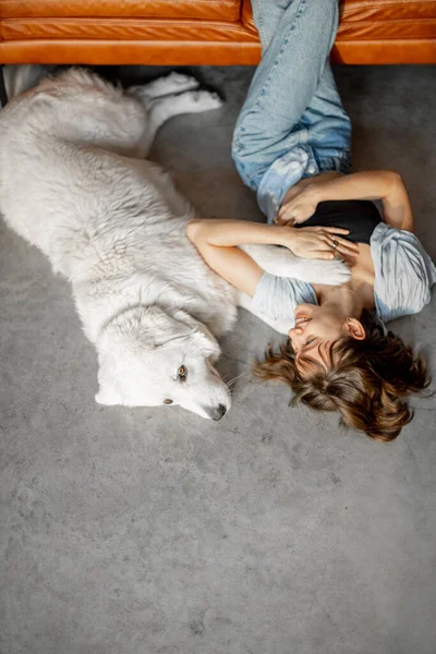 Woman lying relaxed with her adorable dog on the floor near couch at home. Concept of home comfort and friendship with pets. Maremma sheepdog with caucasian woman
