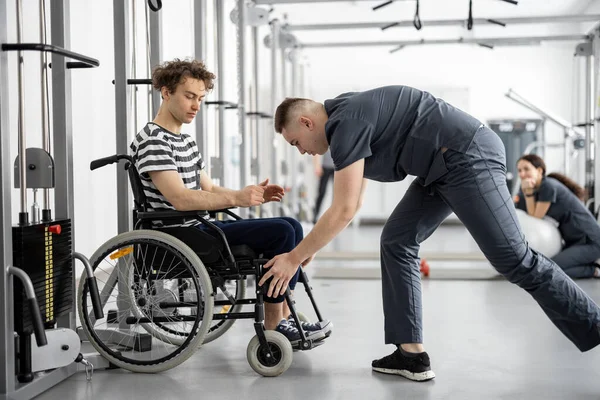 Rehabilitation specialist helps a guy in a wheelchair to do exercise on decompression simulator for recovery from injury. Concept of physical therapy for people with disabilities