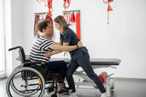 Nurse of the rehabilitation center helps man to get up from a wheelchair at clinic. Concept of medical care for people with disabilities and physical rehabilitation