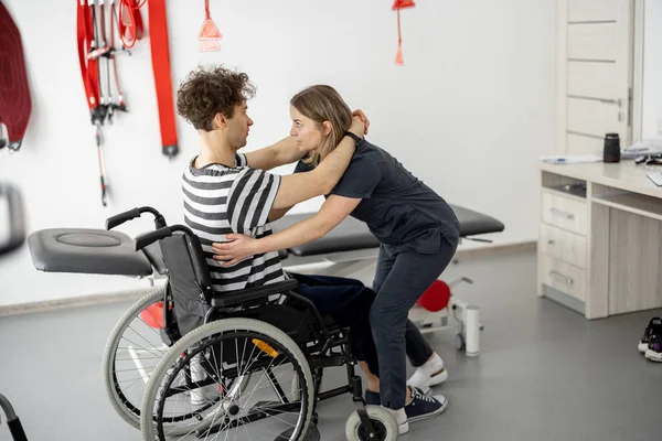 Nurse of the rehabilitation center helps man to get up from a wheelchair at clinic. Concept of medical care for people with disabilities and physical rehabilitation