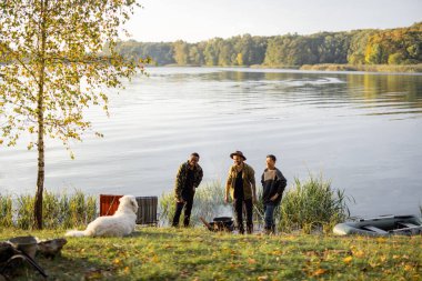 Landscape of beautiful lake with group of male friends having a picnic while fishing on the coast. Leisure, weekend and vacation in nature